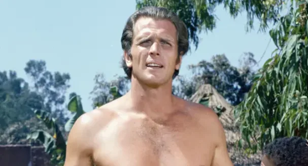 A shirtless man with dark hair and a muscular build stands outdoors under a bright sky, looking upward with a serious…