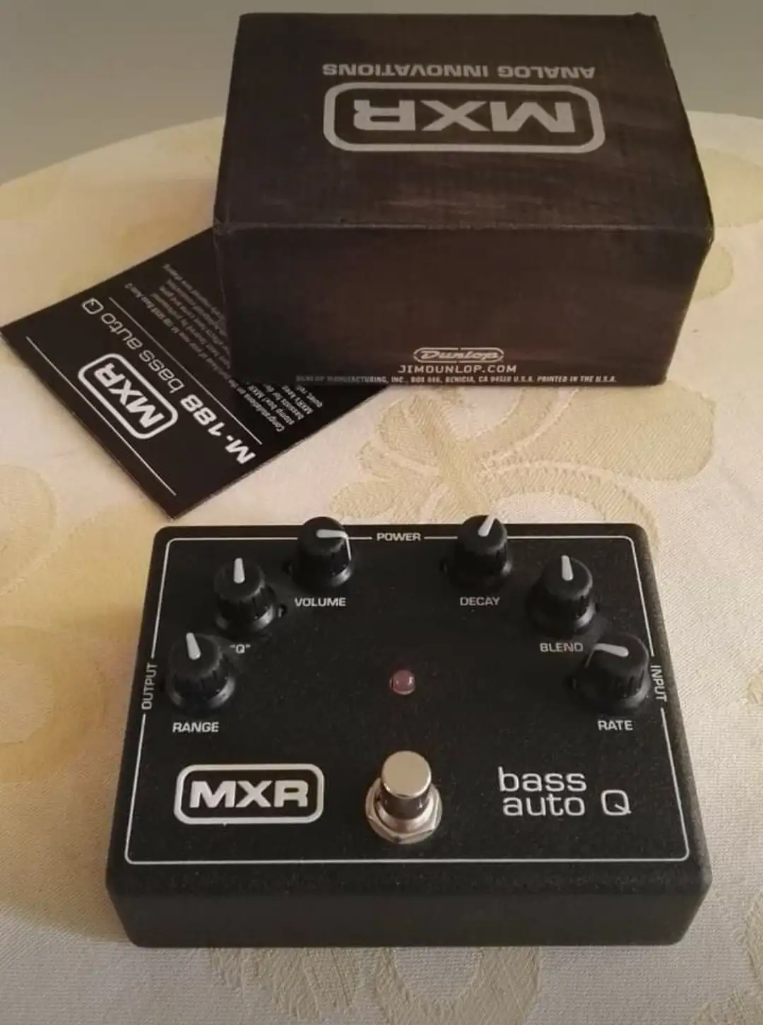 A black MXR M-188 bass auto Q effects pedal sits on a patterned tablecloth, with its original black box and instruction…