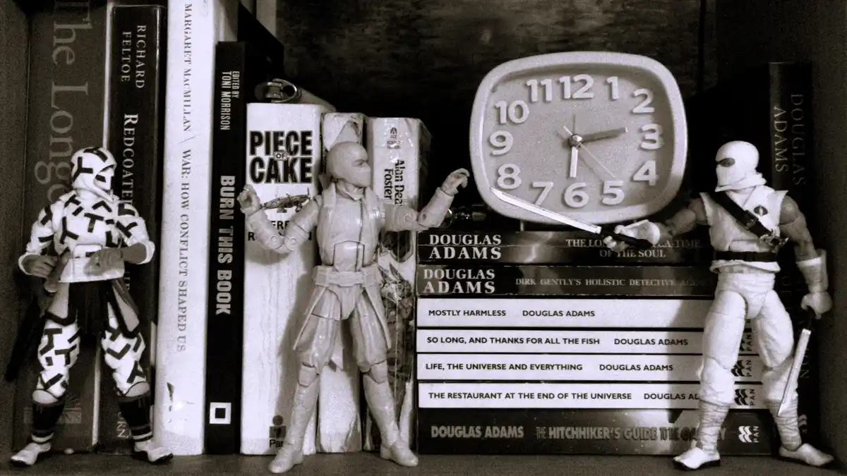 A sepia-toned photo shows three ninja action figures standing around a stack of Douglas Adams books, with one figure holding…