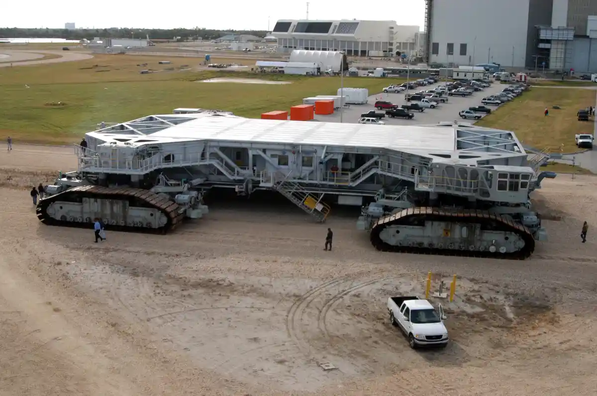 The image shows a massive tracked vehicle with a building-like structure on top, parked on a gravel lot near a parking lot…