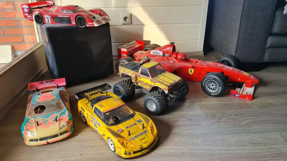 A collection of five remote-controlled toy cars and trucks of various types and colors are arranged on a wooden floor near a…