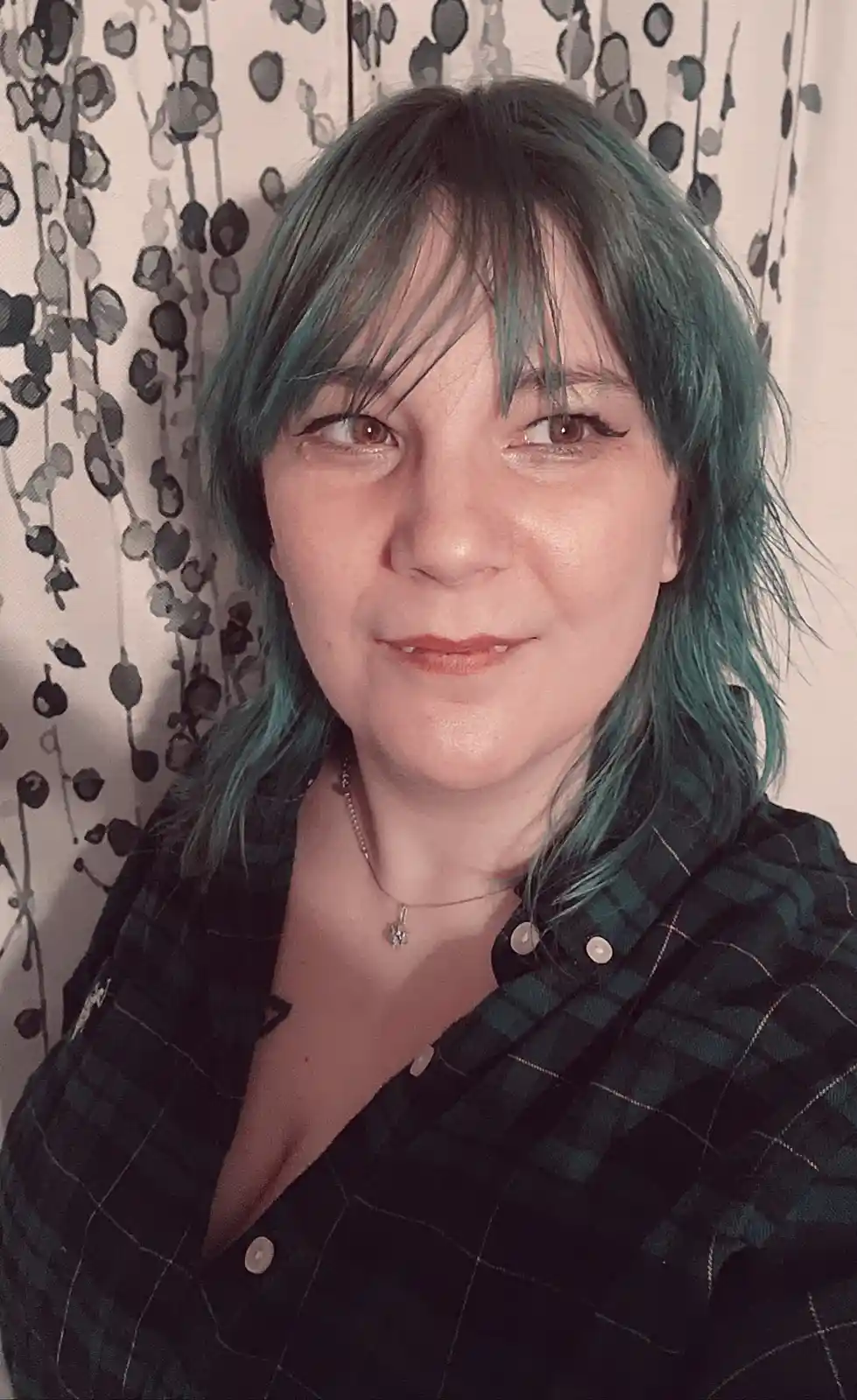 A person with teal-green hair and bangs looks directly at the camera with a slight smile while wearing a dark plaid shirt…