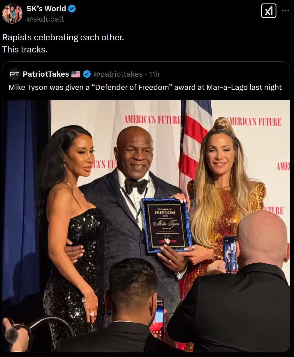 A man in a suit holds a 'Defender of Freedom' award plaque while standing between two women in formal dresses at an event…