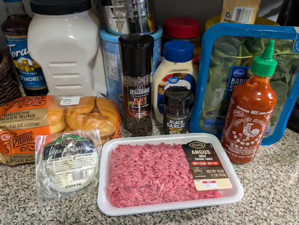 A kitchen counter displays ingredients for a burger including ground beef, brioche buns, mozzarella cheese, sriracha sauce…