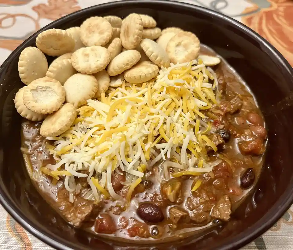 A close-up photo of a dark brown bowl filled with chili, topped with a generous pile of shredded yellow and white cheese and…