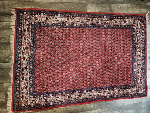An overhead photo shows a rectangular, ornate rug with a red field and intricate blue, white, and burgundy border patterns…