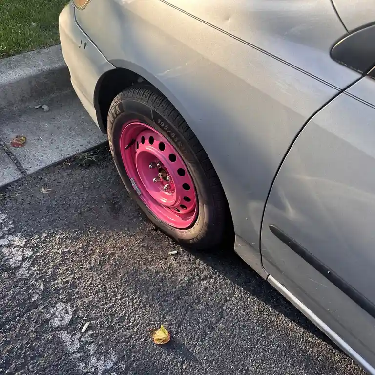 A close-up photo shows the rear wheel of a silver car parked on asphalt, featuring a bright pink aftermarket rim with…