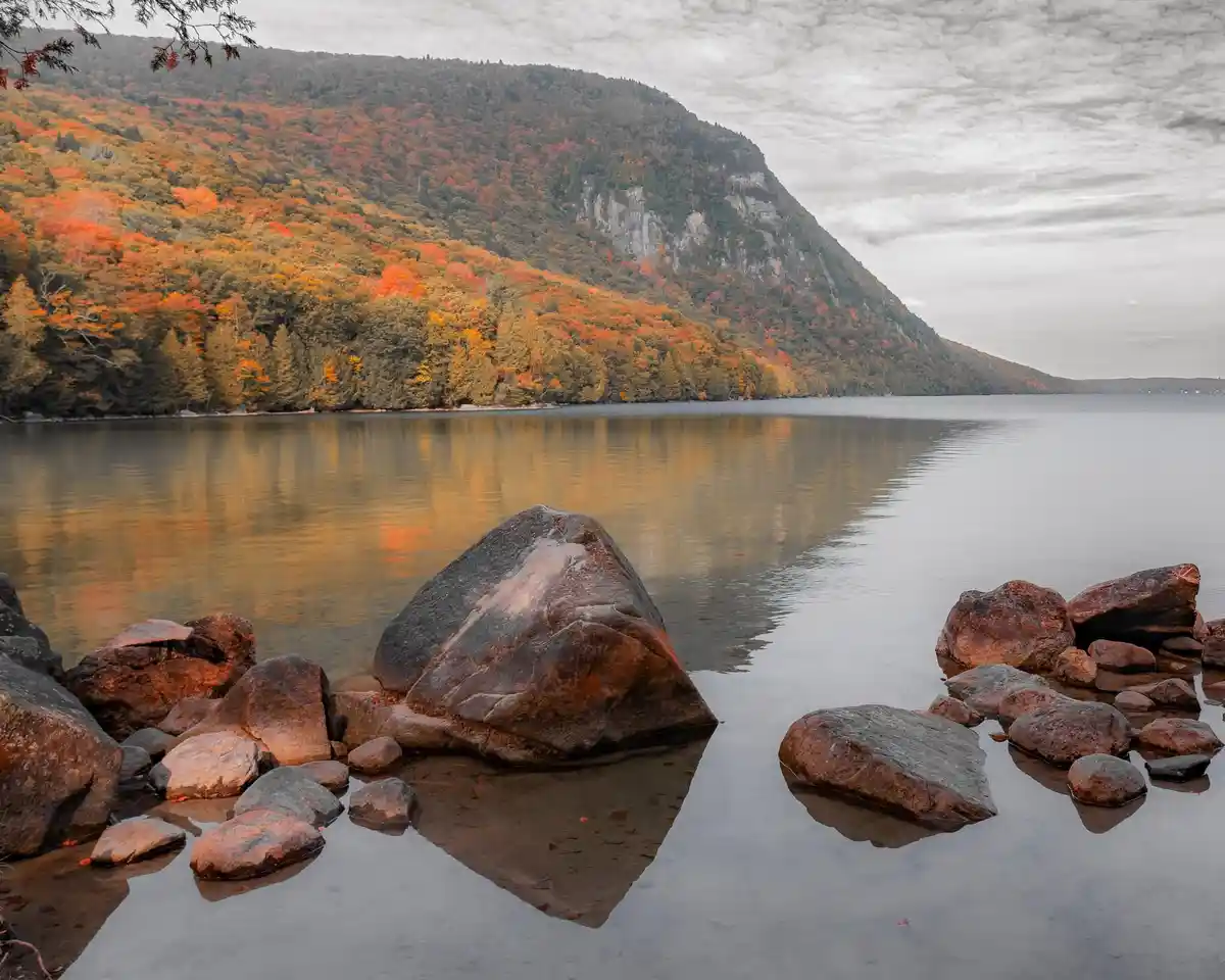 A serene autumn landscape featuring a calm lake reflecting a forested hillside with vibrant orange and red foliage under an…