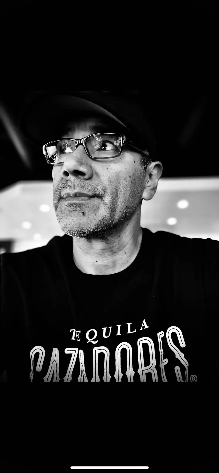 A black-and-white selfie of a man wearing glasses and a black cap, looking upward with a slight smirk. He wears a black…