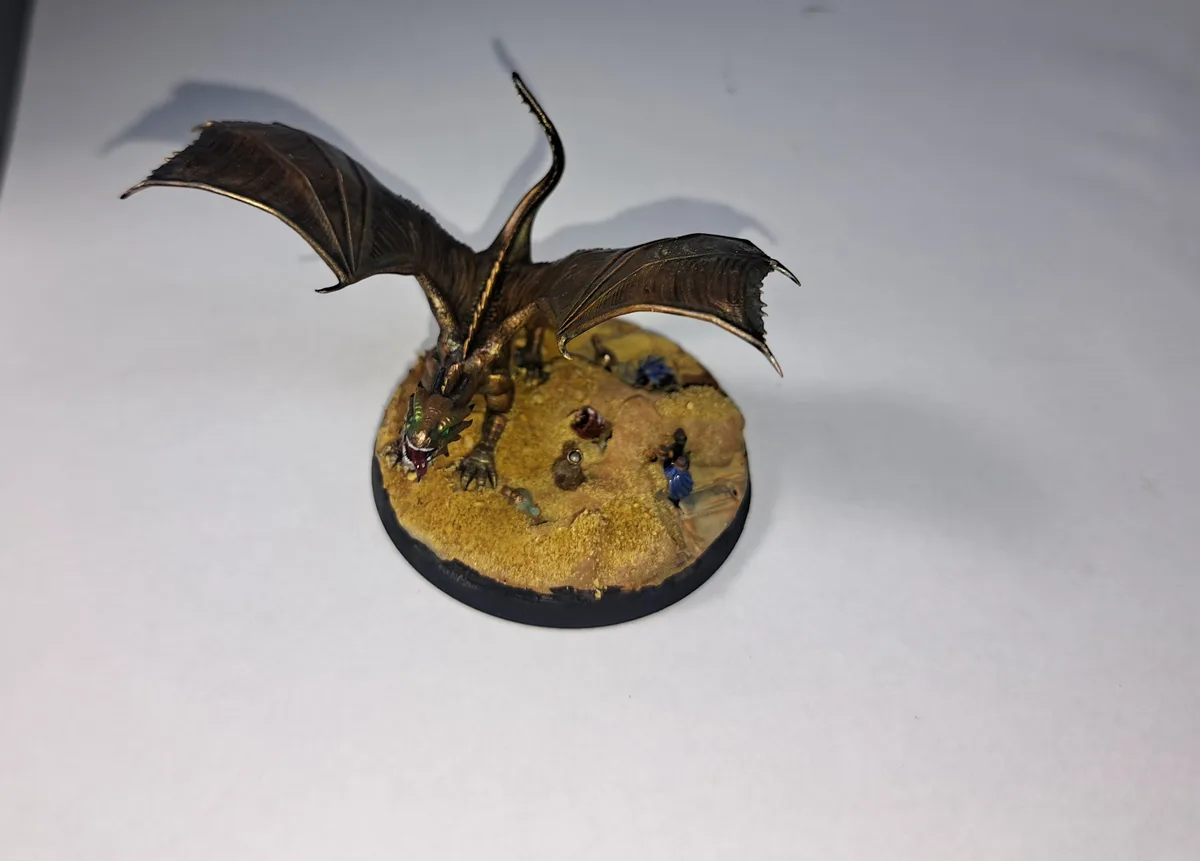 A painted miniature dragon with spread wings and an open mouth stands on a circular base depicting a sandy terrain with tiny…