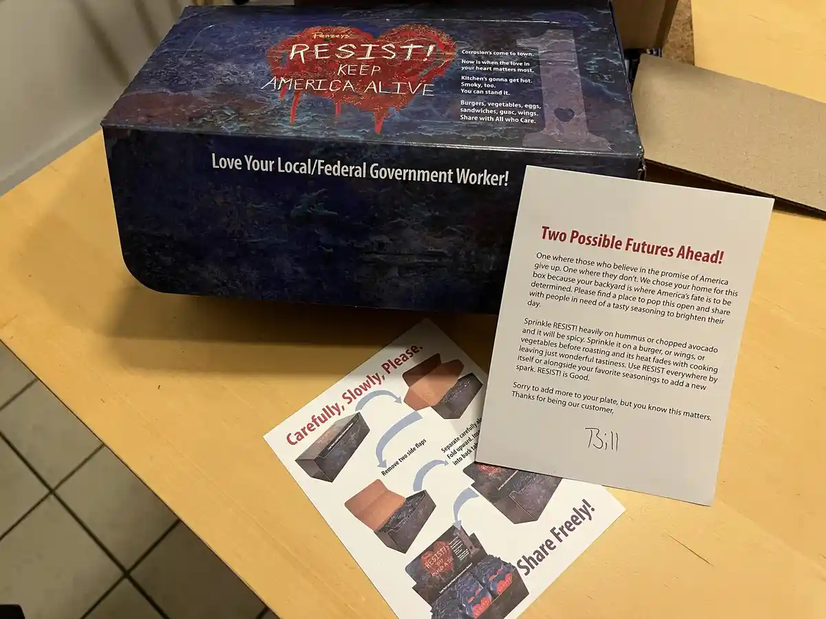 A dark blue box labeled 'RESIST! KEEP AMERICA ALIVE' with a red heart graphic sits on a wooden table, accompanied by a card…