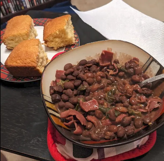A close-up photo shows a bowl of dark beans mixed with bacon pieces and green peppers, sitting on a red and white patterned…