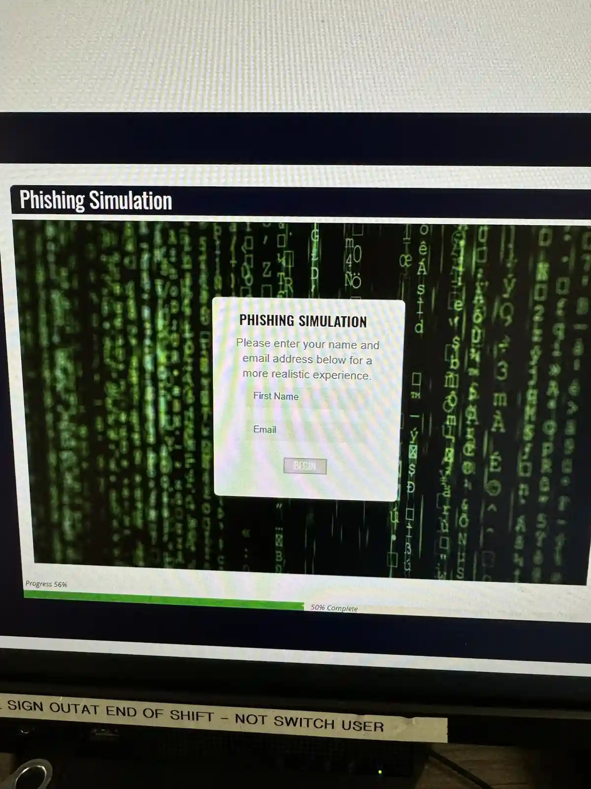 A computer monitor displays a phishing simulation webpage with a Matrix-style green code background. A pop-up window titled…