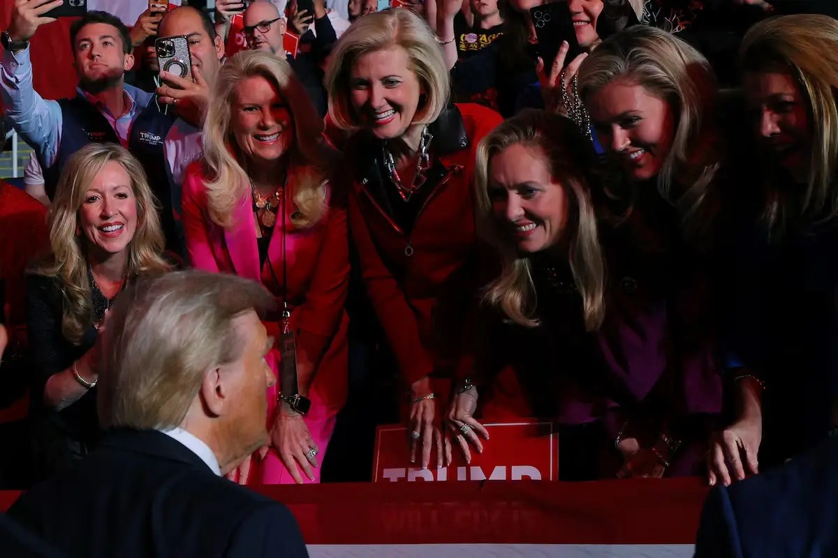 A photo shows Donald Trump from behind, facing a group of smiling women leaning over a red table with his name on it. The…