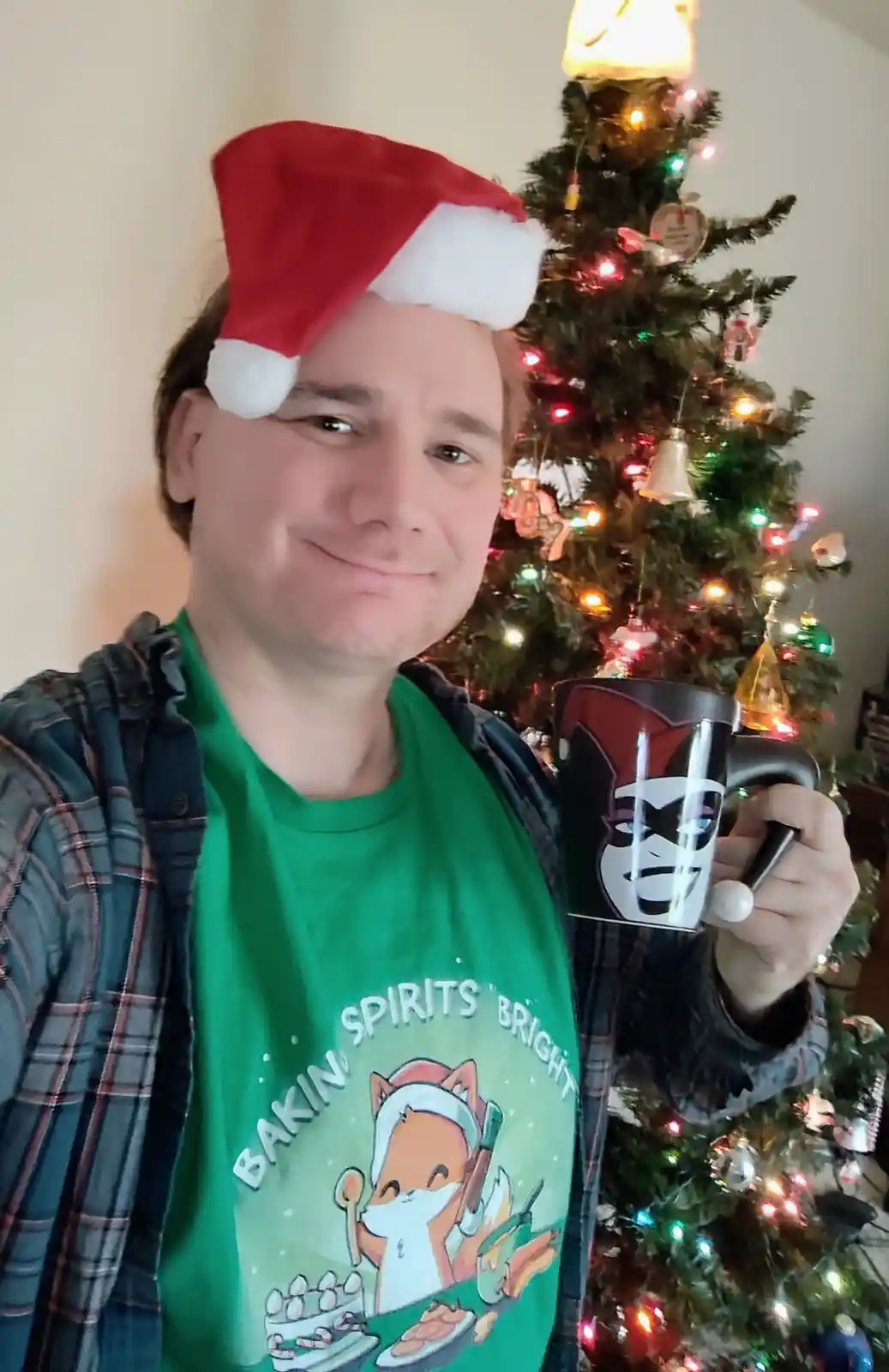 A man wearing a red and white Santa hat and a green t-shirt with a cartoon fox baking and the text 'BAKIN SPIRITS BRIGHT'…