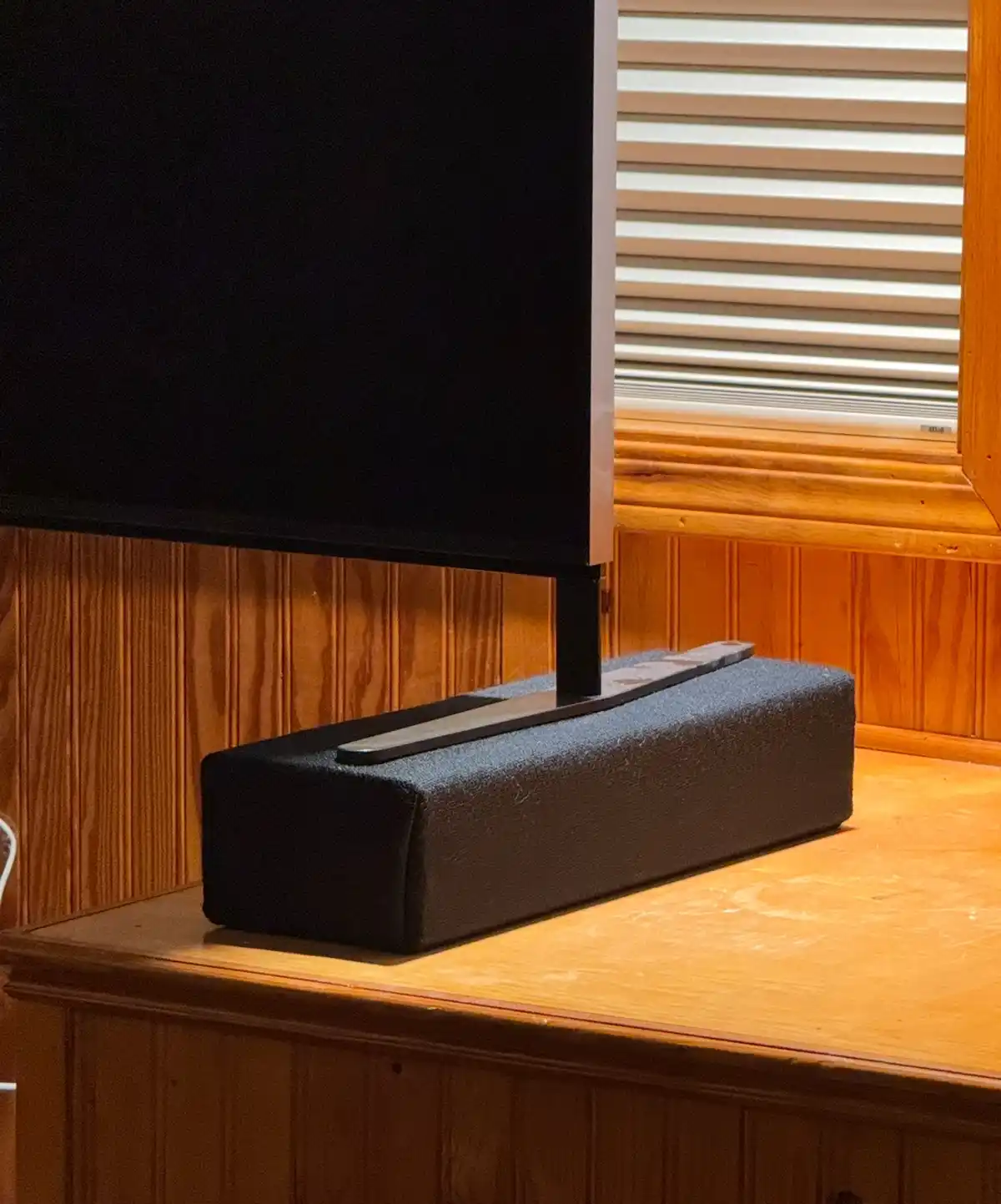 A black rectangular speaker sits on a wooden TV stand beneath a large TV, with a window and blinds visible in the…