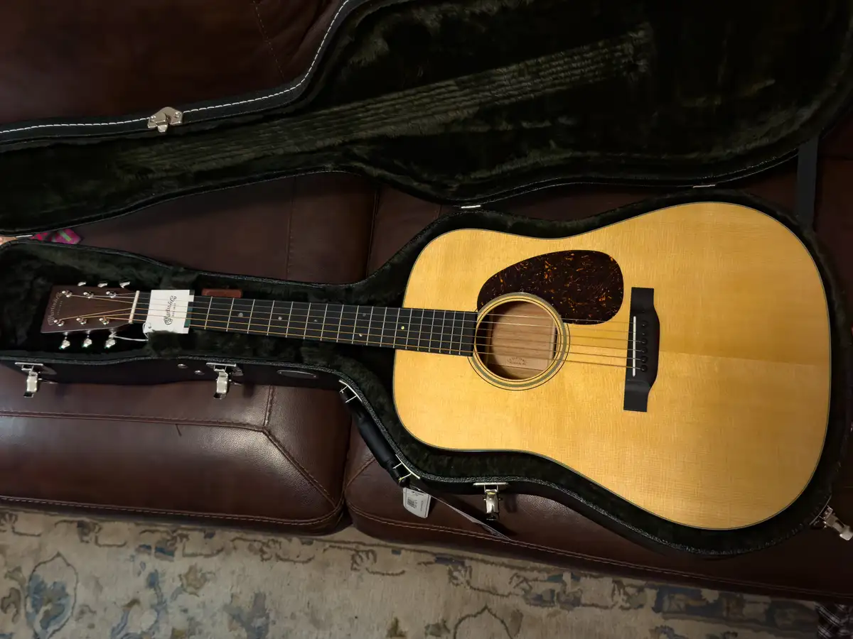 An acoustic guitar with a natural wood finish and black pickguard rests inside an open, black, plush-lined hard case. The…