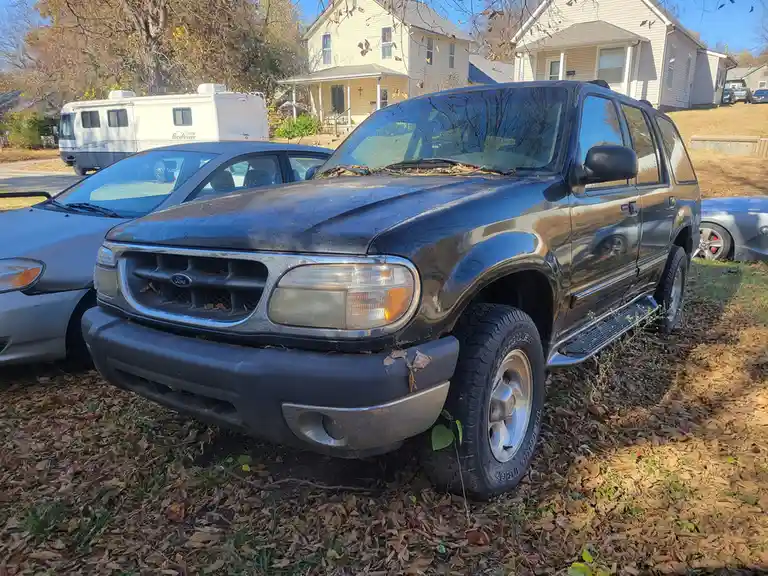 A black Ford Explorer SUV is parked on a grassy area covered in fallen autumn leaves, with other cars and houses visible in…