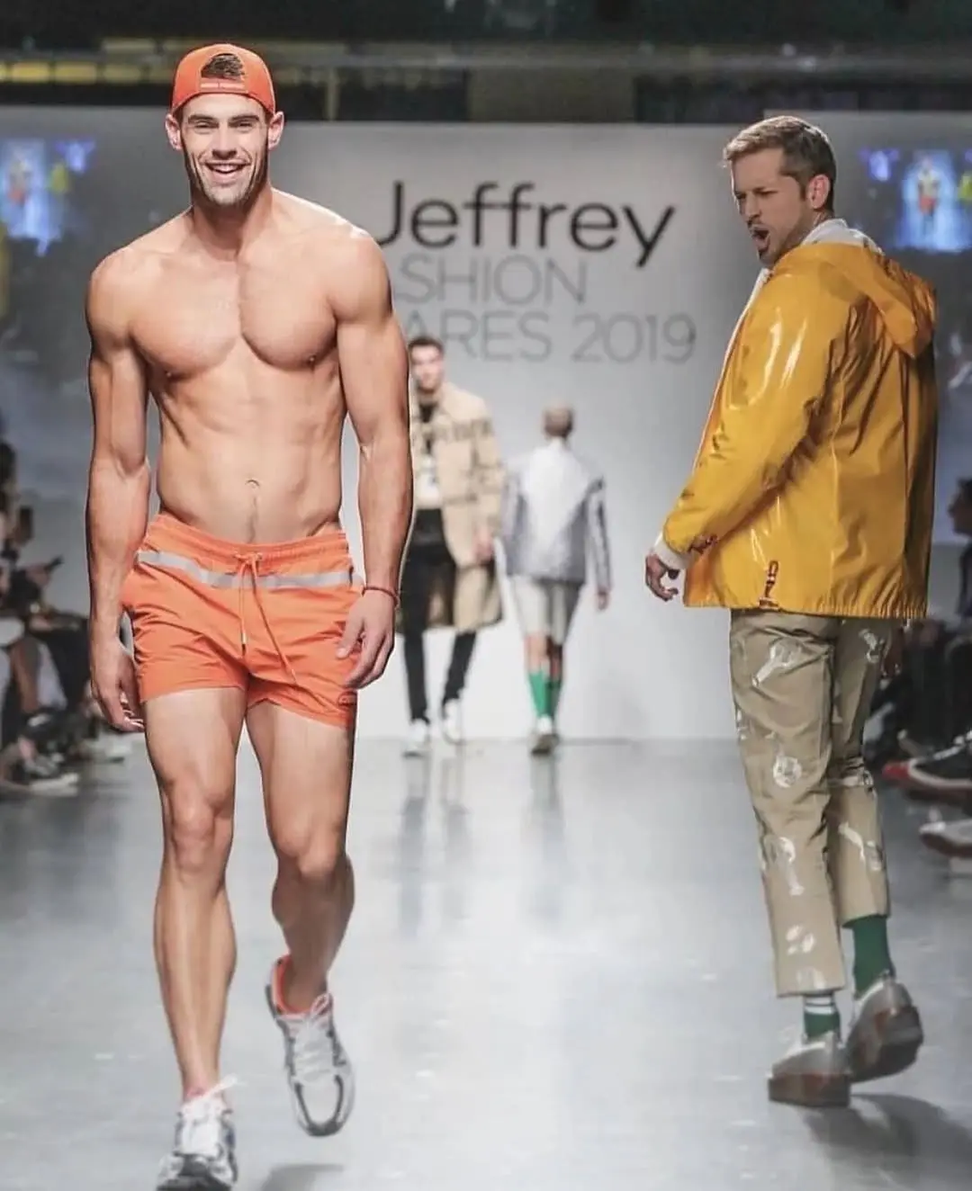 A muscular male model in orange swim trunks and cap walks confidently down a runway at 'Jeffrey Fashion Ares 2019', while…