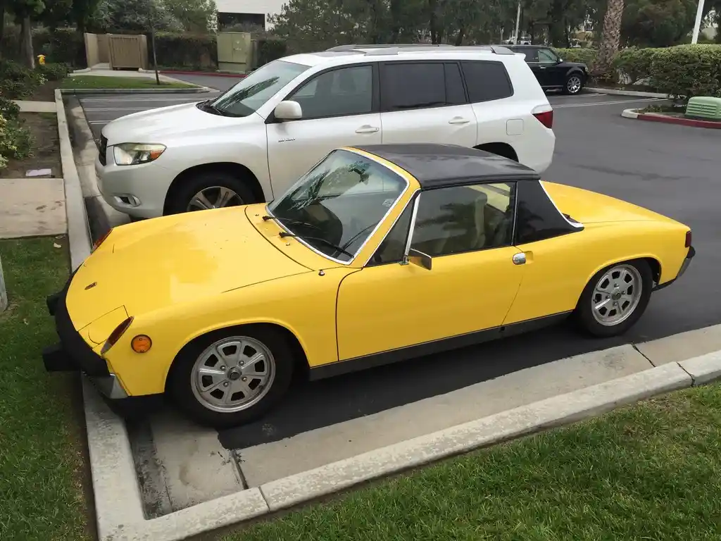 A bright yellow vintage Porsche 914 with a black soft top is parked in a parking space next to a white SUV. The car is…