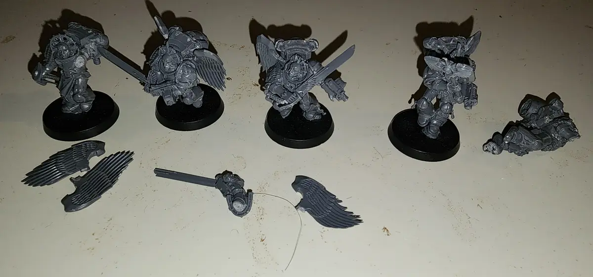 Four grey miniature figures in armor stand on black bases, while scattered around them are detached wings, a sword, and a…