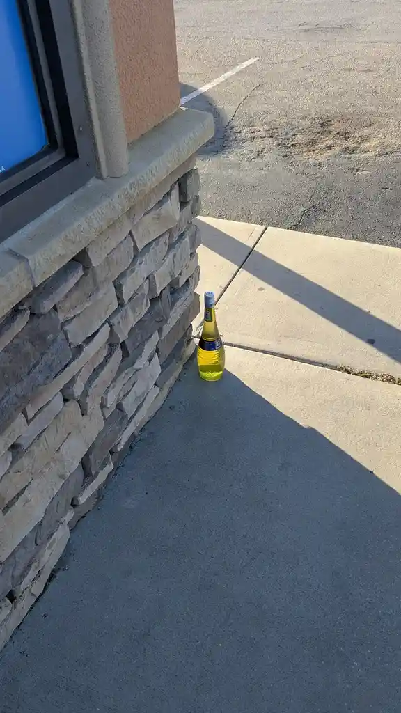 A single bottle of yellow liquid, likely wine, sits upright on a concrete sidewalk next to a stone-clad building wall. The…