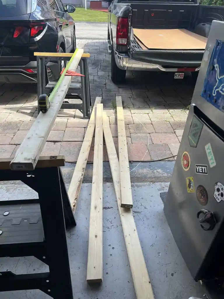 An outdoor driveway scene with several wooden planks, some resting on sawhorses with a measuring tape and speed square on…