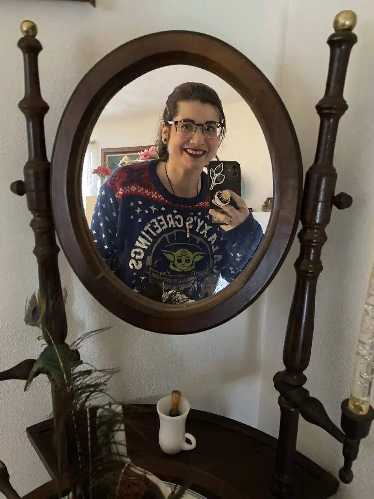 A person with glasses and dark lipstick smiles while taking a mirror selfie, wearing a blue sweater with a Yoda graphic and…