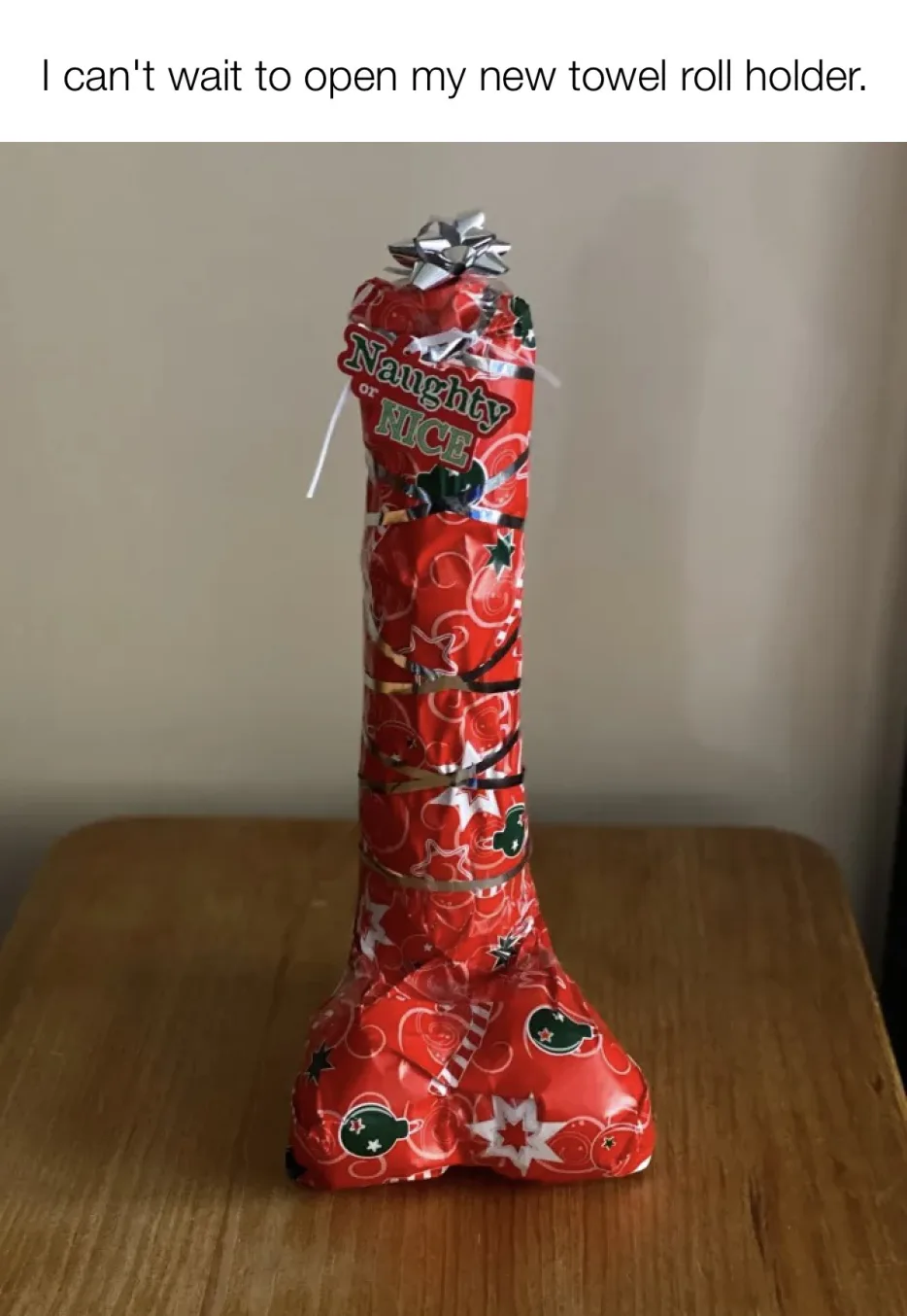 A tall, red Christmas-themed gift-wrapped object, resembling a towel roll holder, sits on a wooden table. It's wrapped in…