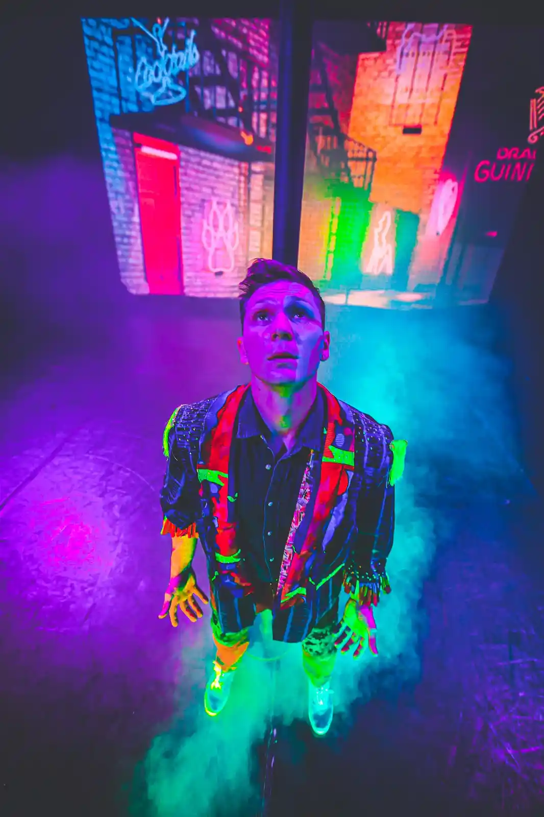 A man in a colorful, neon-lit nightclub setting with a surprised or overwhelmed expression, standing in smoke with vibrant…