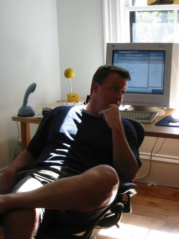 a man sits back in a chair at a desk with a computer monitor, keyboard, and lamp, looking thoughtful with his hand on his…