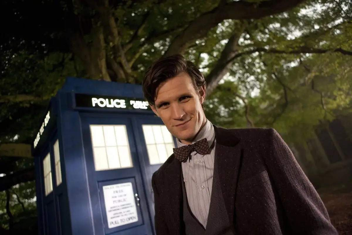A man in a brown suit and bow tie stands smiling in front of a blue police box at night, with trees in the background. The…