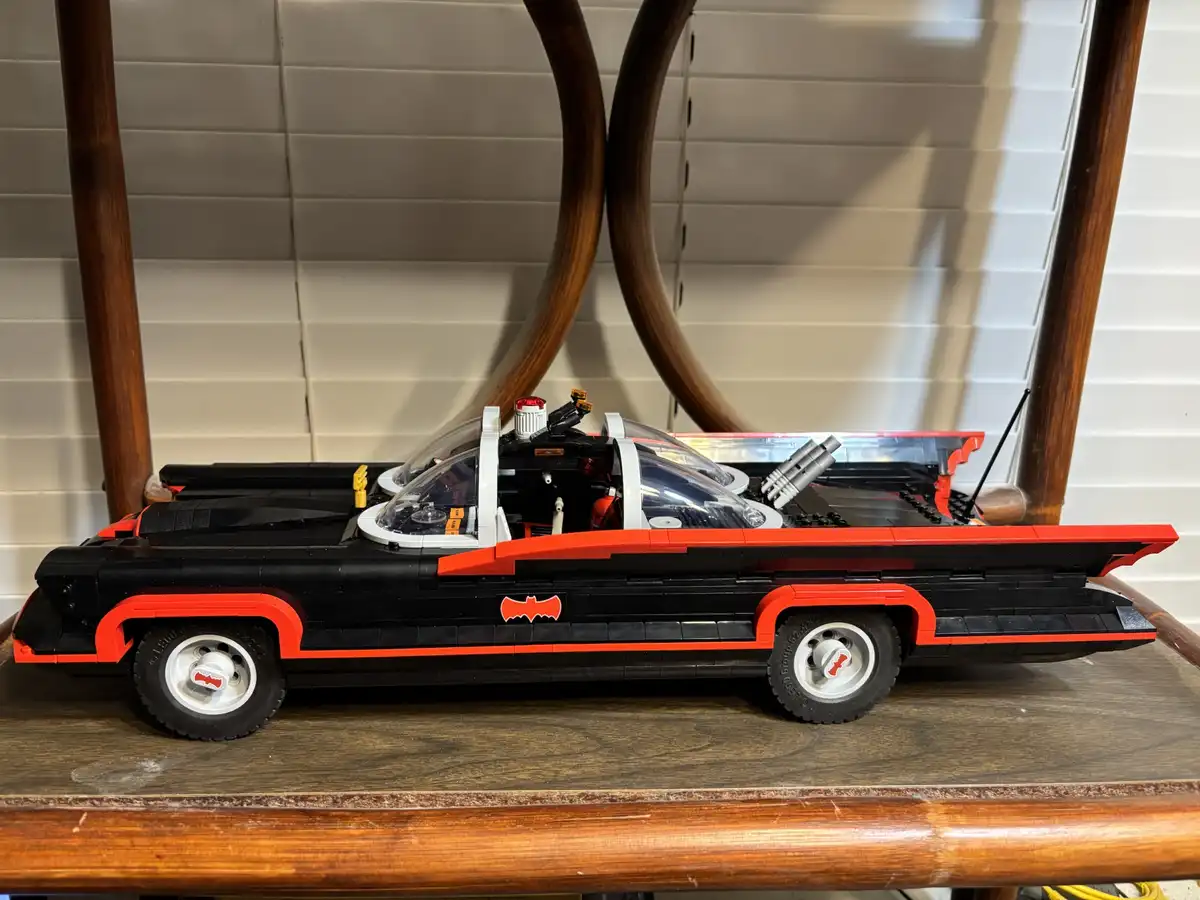 A detailed LEGO model of the classic 1960s Batmobile, black with red trim and a bat symbol, sits on a wooden shelf. The…
