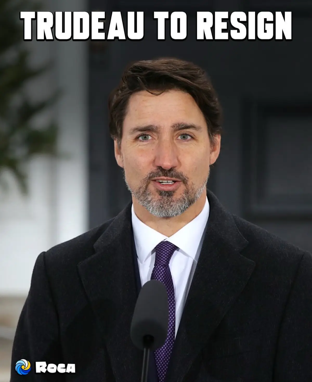 A close-up photo of a man with a beard and mustache wearing a dark suit, white shirt, and purple tie, standing in front of a…