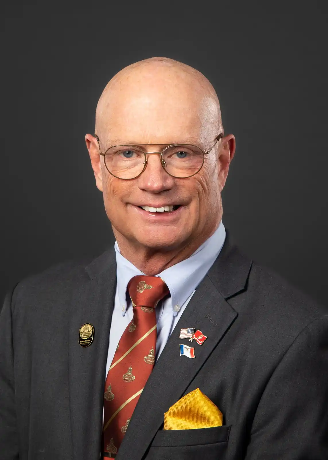 A bald man with glasses, wearing a dark suit, light blue shirt, and red tie with gold patterns, smiles at the camera. He has…