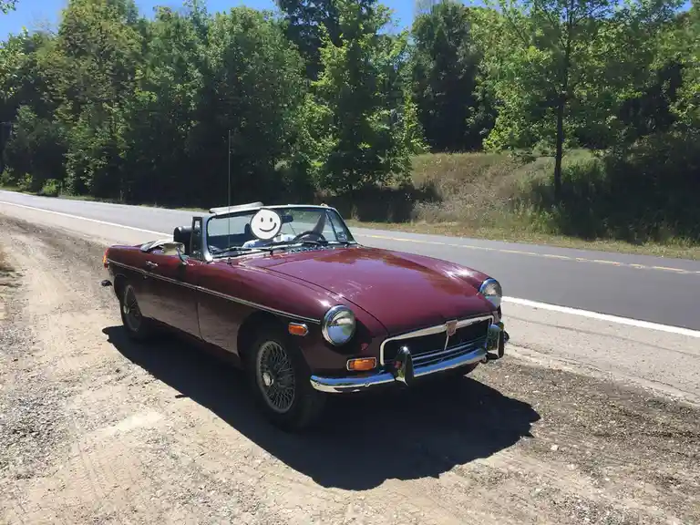 A maroon classic convertible car, likely an MG MGB, is parked on a dirt shoulder next to a paved road with trees in the…