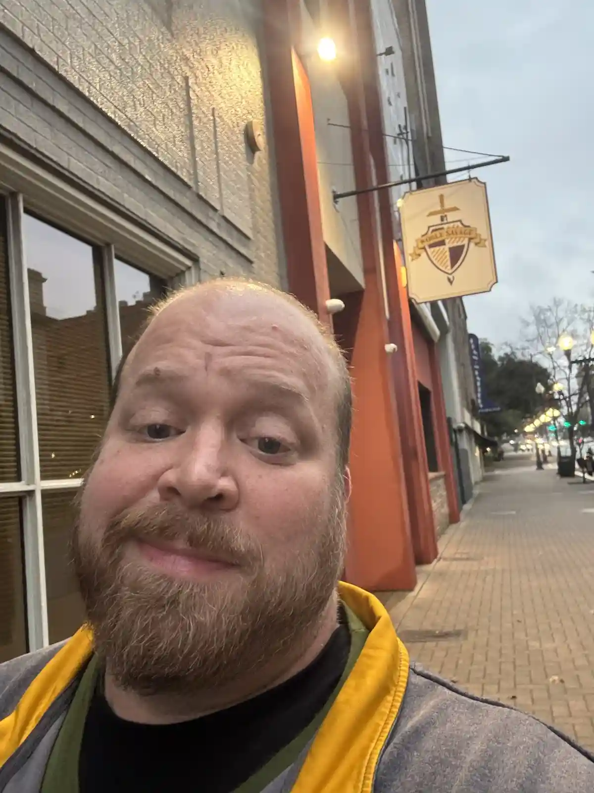 A man with a beard and a gray jacket with yellow trim takes a selfie outside a building with a 'Noble Savage' sign. He has a…