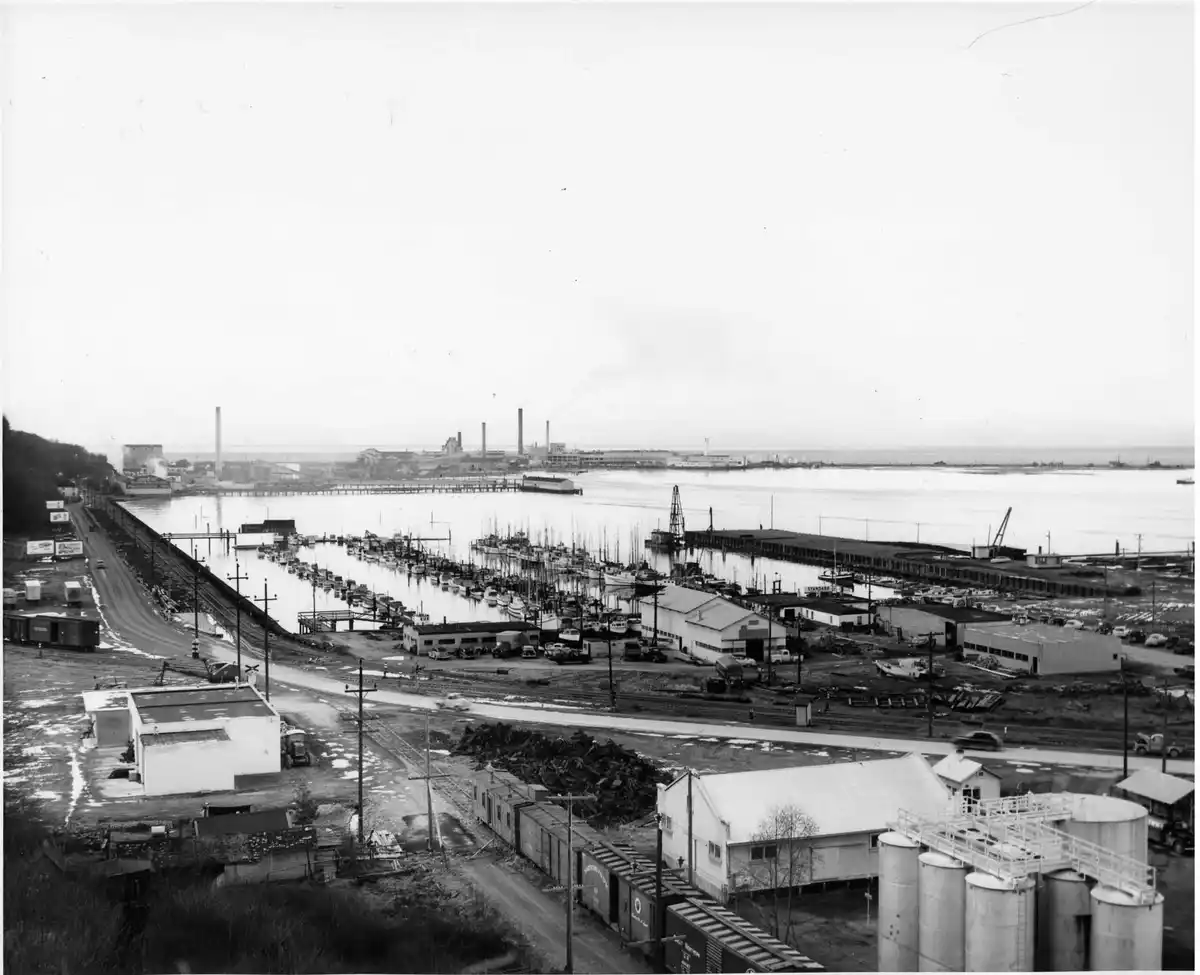 A black-and-white historical photo shows a busy industrial harbor with docks, boats, factories, and rail lines. Overlaid…