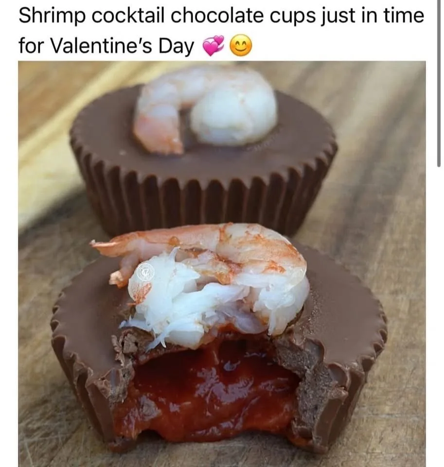 Two chocolate cups sit on a wooden surface, one whole with a cooked shrimp on top, the other broken open to reveal a red…