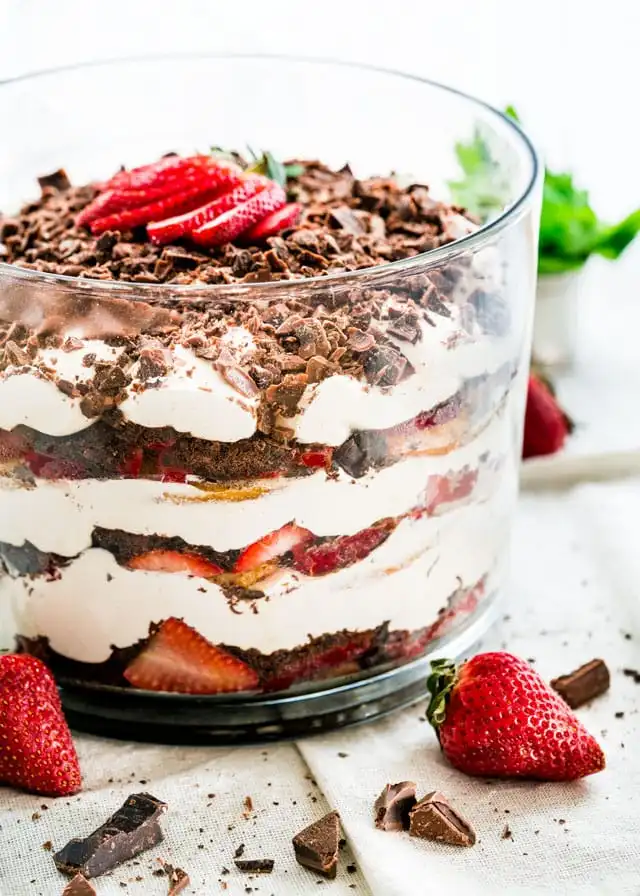 A clear glass trifle bowl contains alternating layers of chocolate cake, whipped cream, and sliced strawberries, topped with…