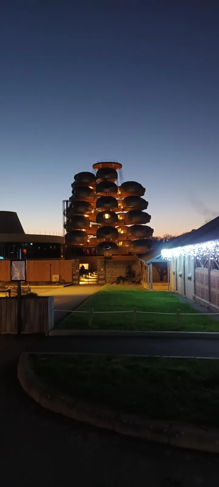 a nighttime photo of a modern, tiered building with glowing, pod-like structures stacked in a tree-like shape, set against a…