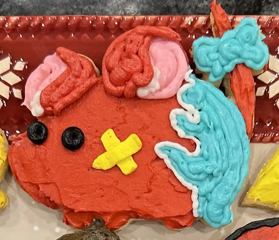 A close-up of a red frosted cookie shaped like a cartoon character with blue frosting hair, blackberry eyes, and a yellow…