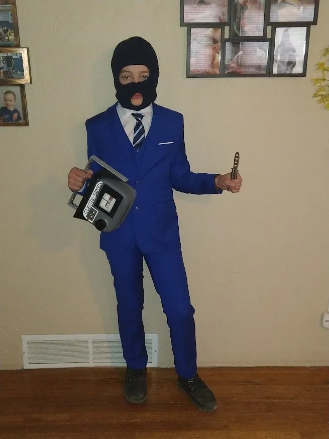 A young person dressed in a bright blue suit, wearing a black ski mask covering their face, holds a large grey boombox in…