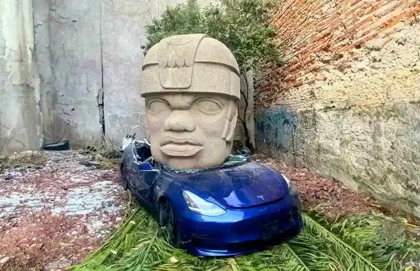 a blue tesla car is parked in an alleyway with a large stone head sculpture placed on its roof, resembling an ancient aztec…