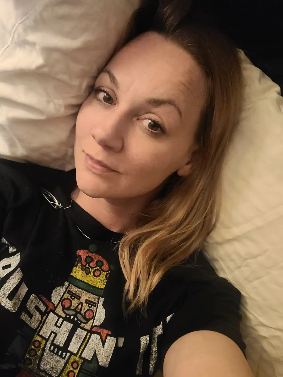 A woman with light brown hair lies on her back on a white pillow, looking directly at the camera with a slightly tired but…