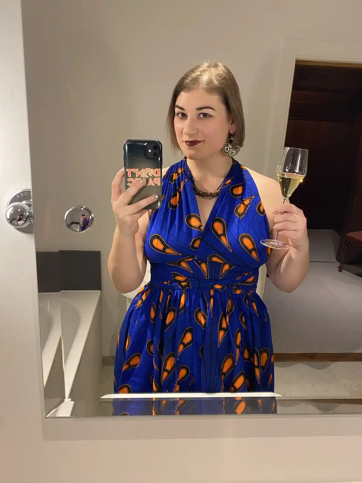 A woman takes a mirror selfie in a bathroom, wearing a blue and orange patterned dress, holding a glass of champagne in one…
