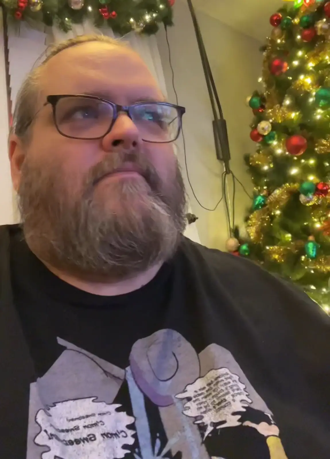 A man with glasses and a beard looks upward with a skeptical expression, standing in front of a decorated christmas tree…