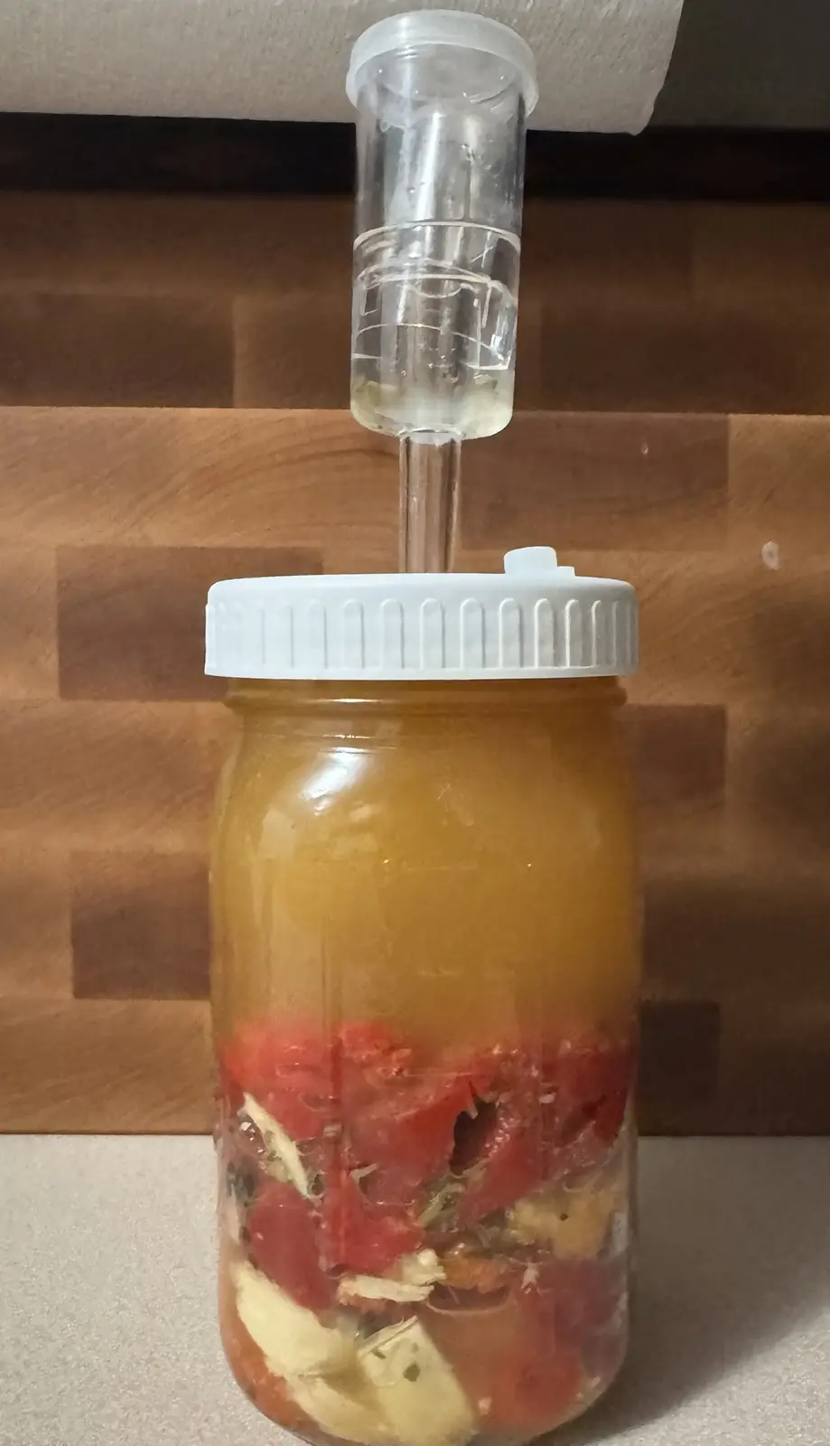 A glass mason jar with a white lid sits on a countertop, filled with chopped red tomatoes and other ingredients submerged in…