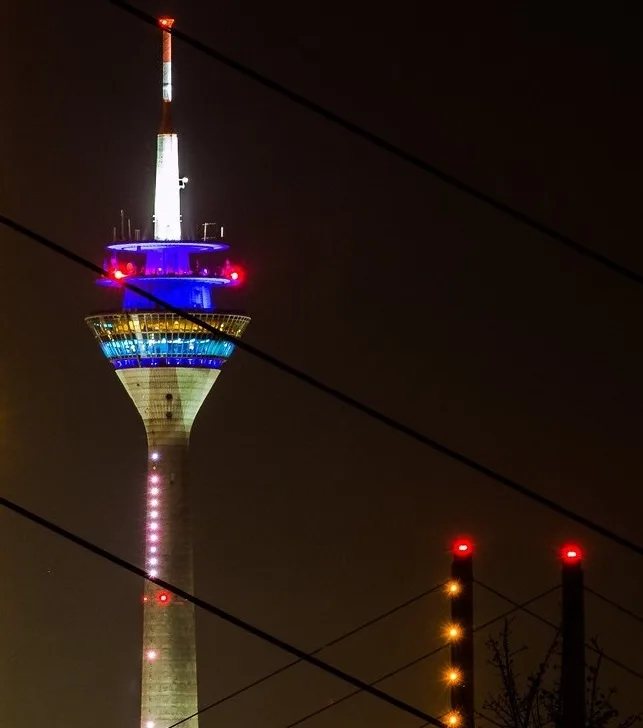 A tall, illuminated tower at night, lit with blue and white lights, with red warning lights on its upper levels and along…