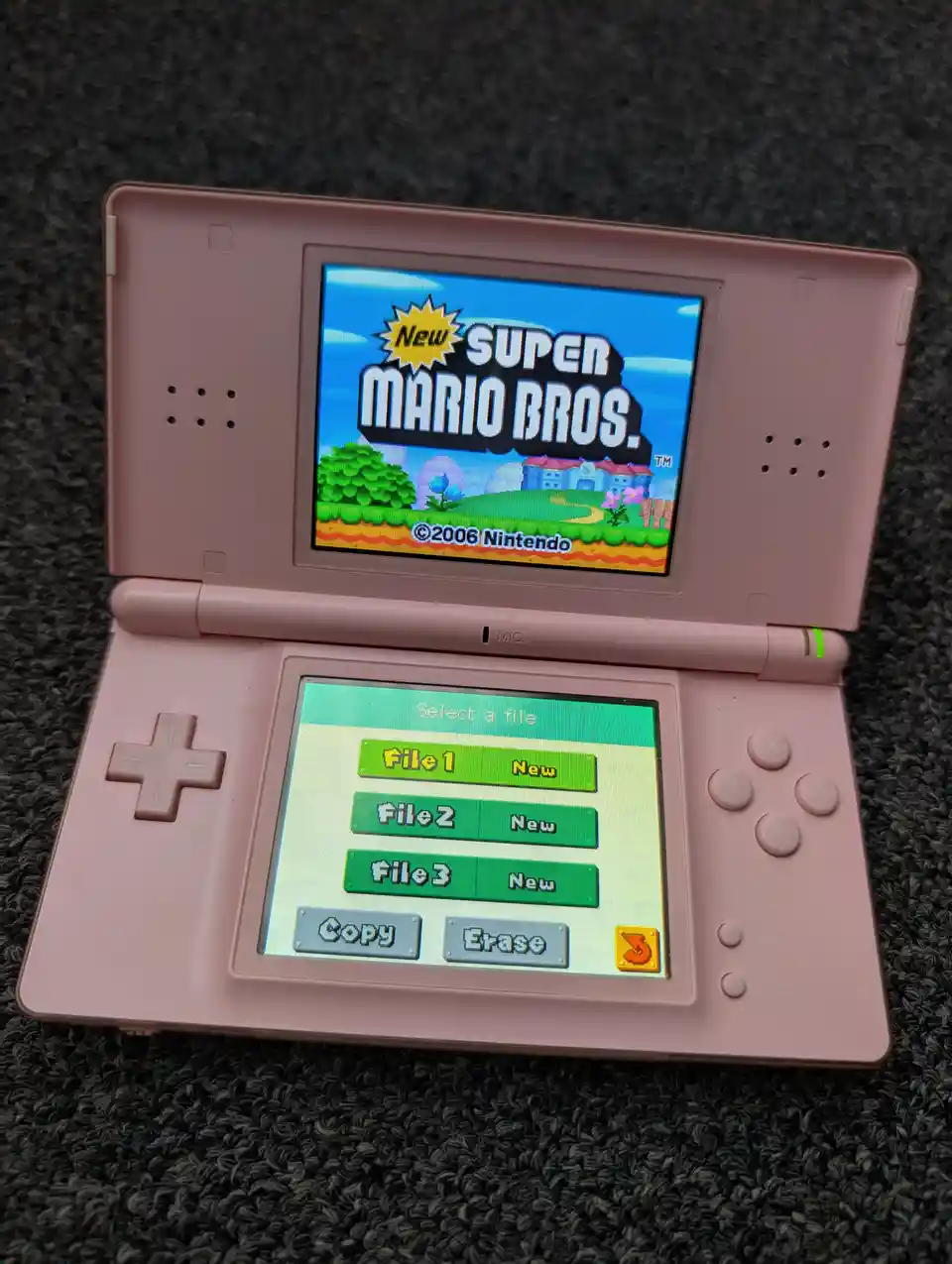 A pink Nintendo DS console displays the title screen of 'New Super Mario Bros.' on its top screen while the bottom screen…